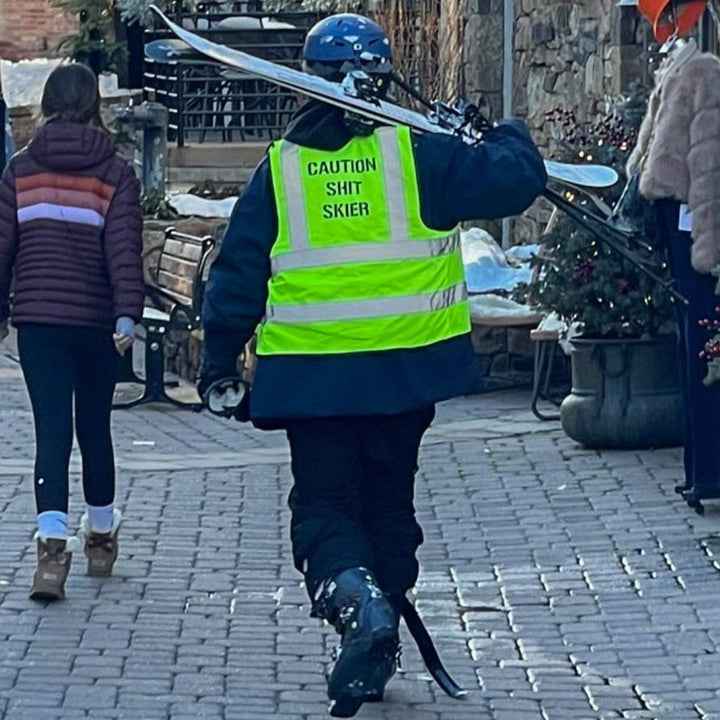 Jerry of the Day Caution Shit Skier Vest, man carrying skis at lodge. Funny ski and snowboard gifts, shirts and apparel. 